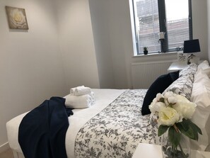 A neatly made bed with a floral bedspread, a dark blue throw blanket, and a vase with white flowers.