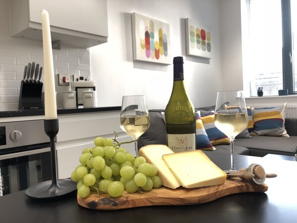 A kitchen counter with a bottle of wine, two glasses of wine, a candle, and a wooden board with grapes and cheese.