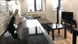 A modern kitchen with a black induction cooktop, a dining area with a black table and chairs, a sofa with colorful cushions, a flat-screen TV, and a clock on the wall. - Cosy One Bedroom Apartment By Creatick (Reading)
