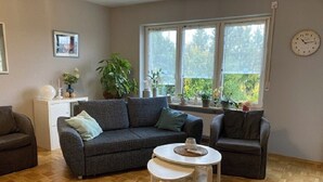 Ein Wohnzimmer mit einer Couch, zwei Sesseln, einem kleinen Tisch und einem Fenster mit Pflanzen.