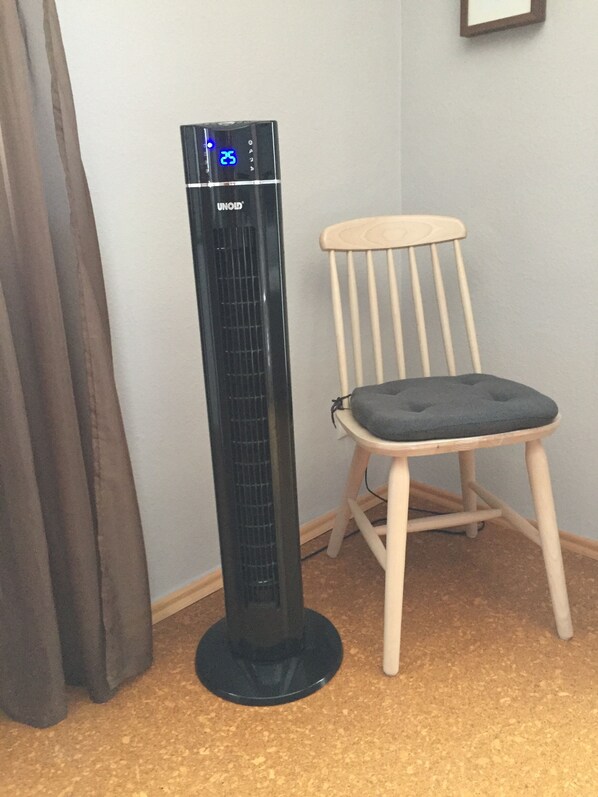 Un ventilador de pie negro con pantalla digital, una silla de madera con cojín y una cortina.