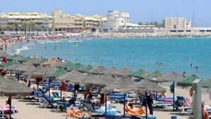 Une plage avec des transats et des parasols, une côte rocheuse et des bâtiments en arrière-plan.