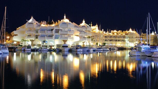 Un port de plaisance où sont amarrés de nombreux bateaux, un grand bâtiment illuminé doté de plusieurs balcons, et une eau calme qui reflète les lumières.