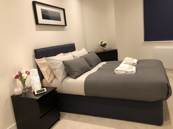 A modern bedroom with a dark headboard, a grey bedspread, and white pillows. There is a bedside table with a clock and a vase of flowers. A framed picture hangs on the wall above the bed.