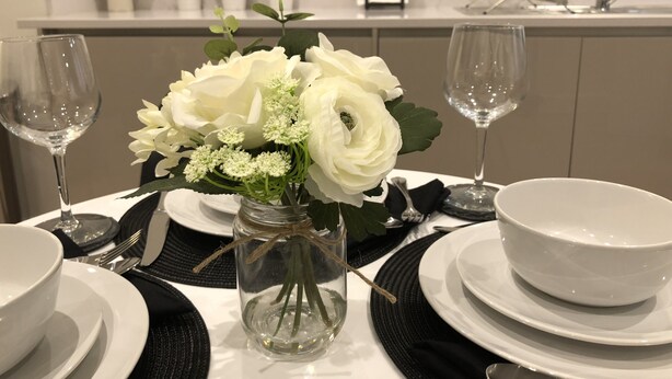A dining table set with white plates, bowls, and cutlery, a clear glass vase with white flowers, and two wine glasses. - Modern Apartment By Creatick (Reading)