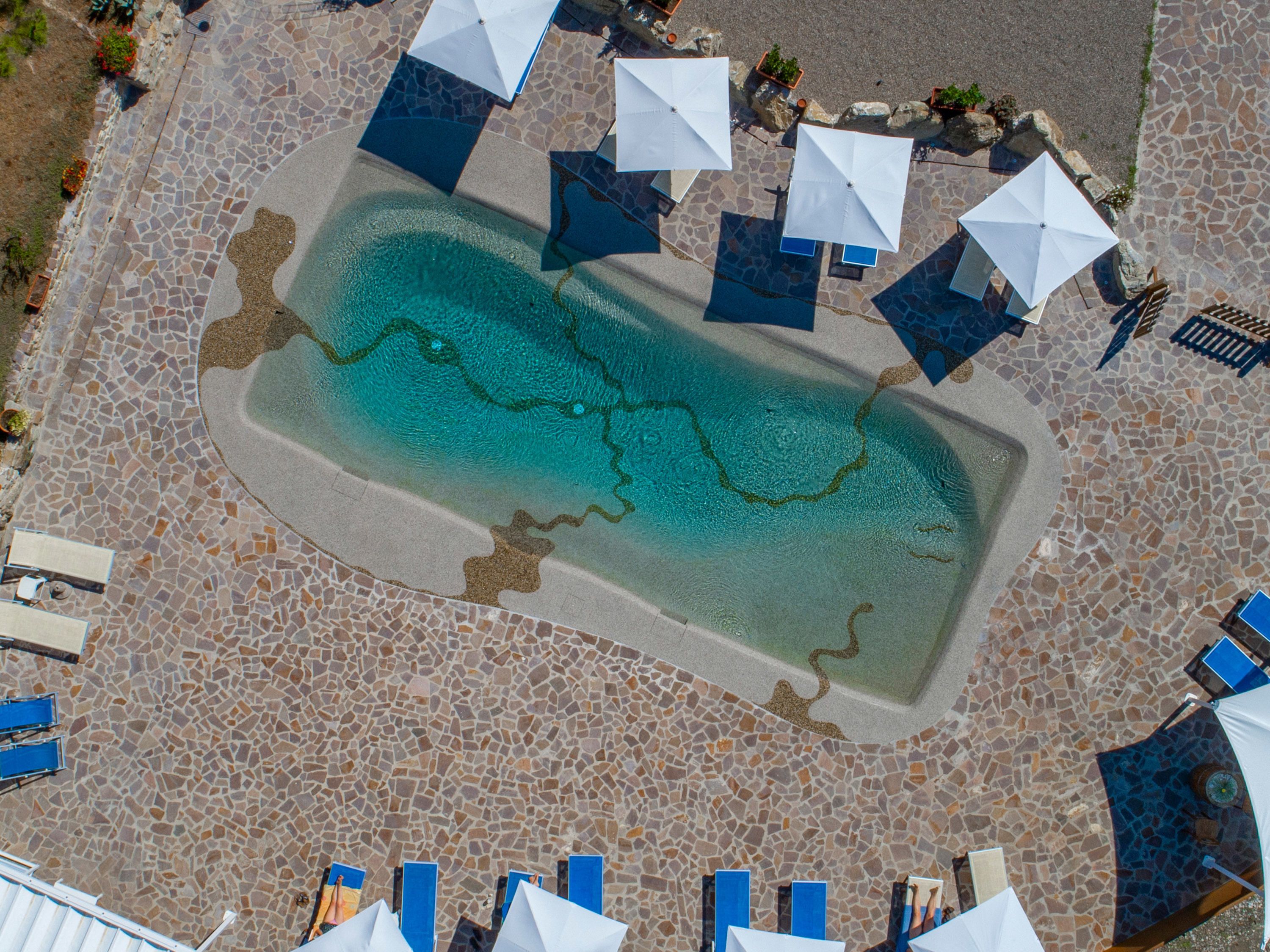 An aerial view of a swimming pool with tiled flooring, surrounded by lounge chairs and umbrellas.