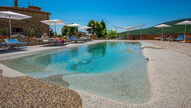 Zona de piscina amb gandules i ombrel·les, envoltada d'edificis de pedra i vegetació.
