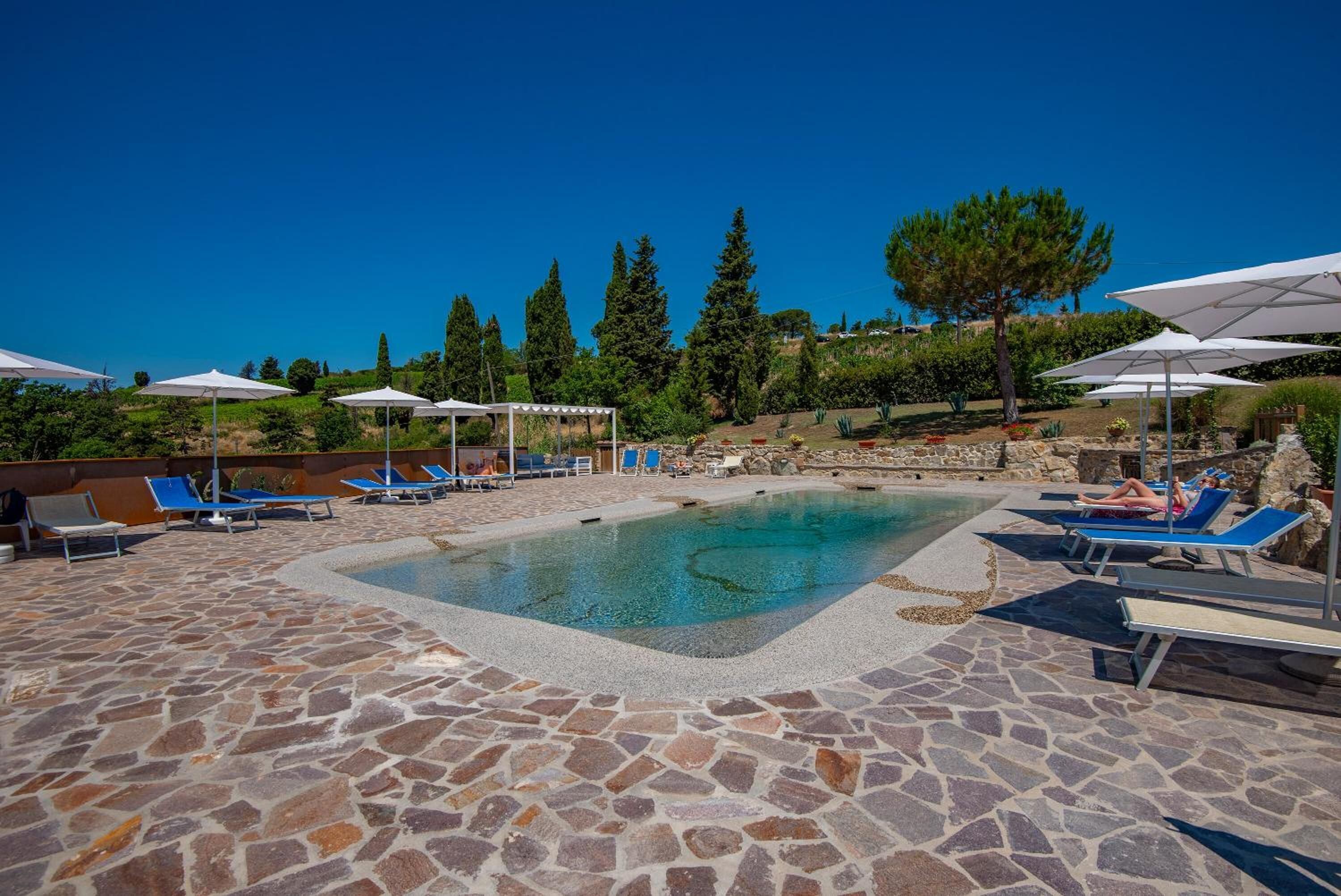 Zona de piscina amb paviment de pedra, gandules i ombrelles.