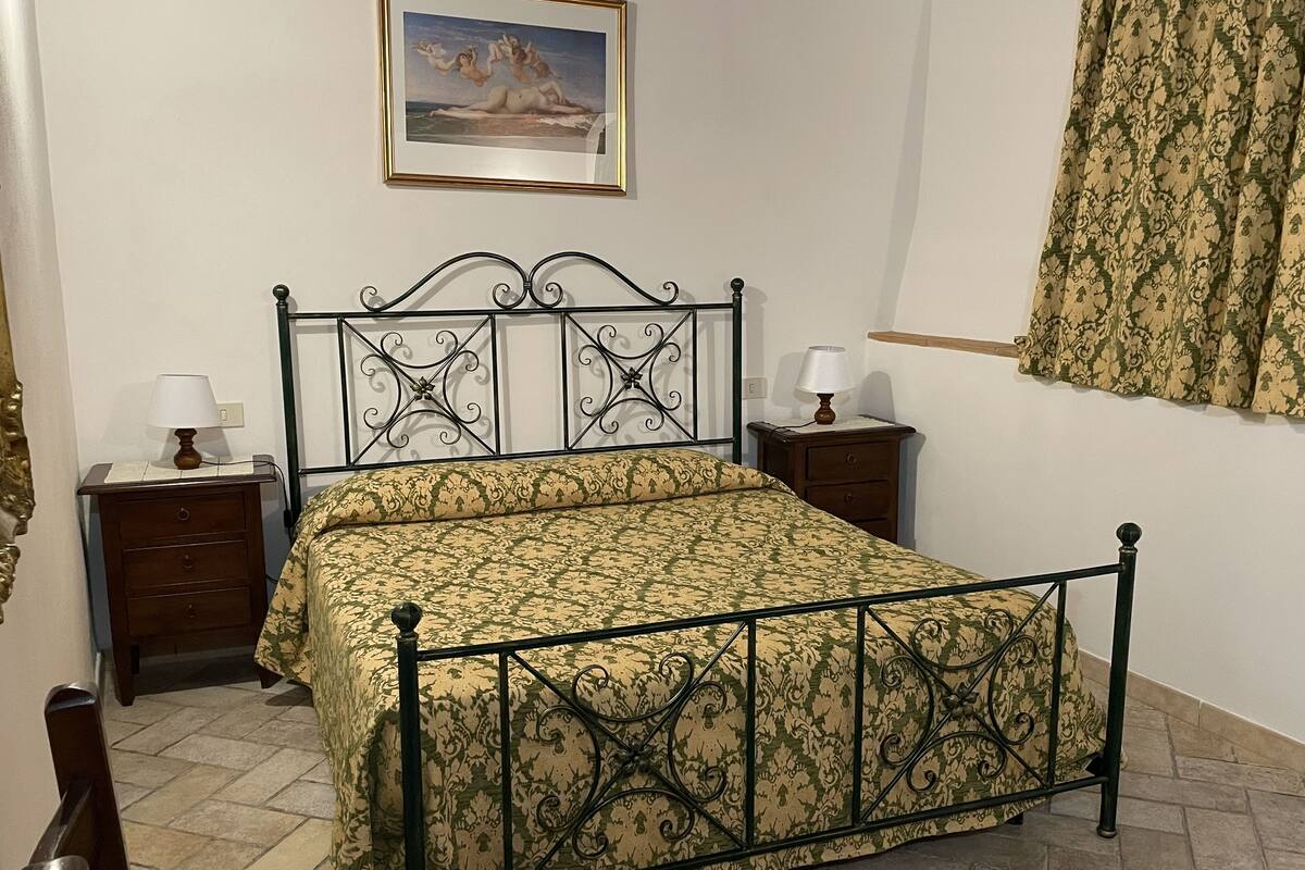 A neatly arranged bedroom with a bed, two bedside tables, a nightstand, a lamp, a framed picture on the wall, and a window with curtains.