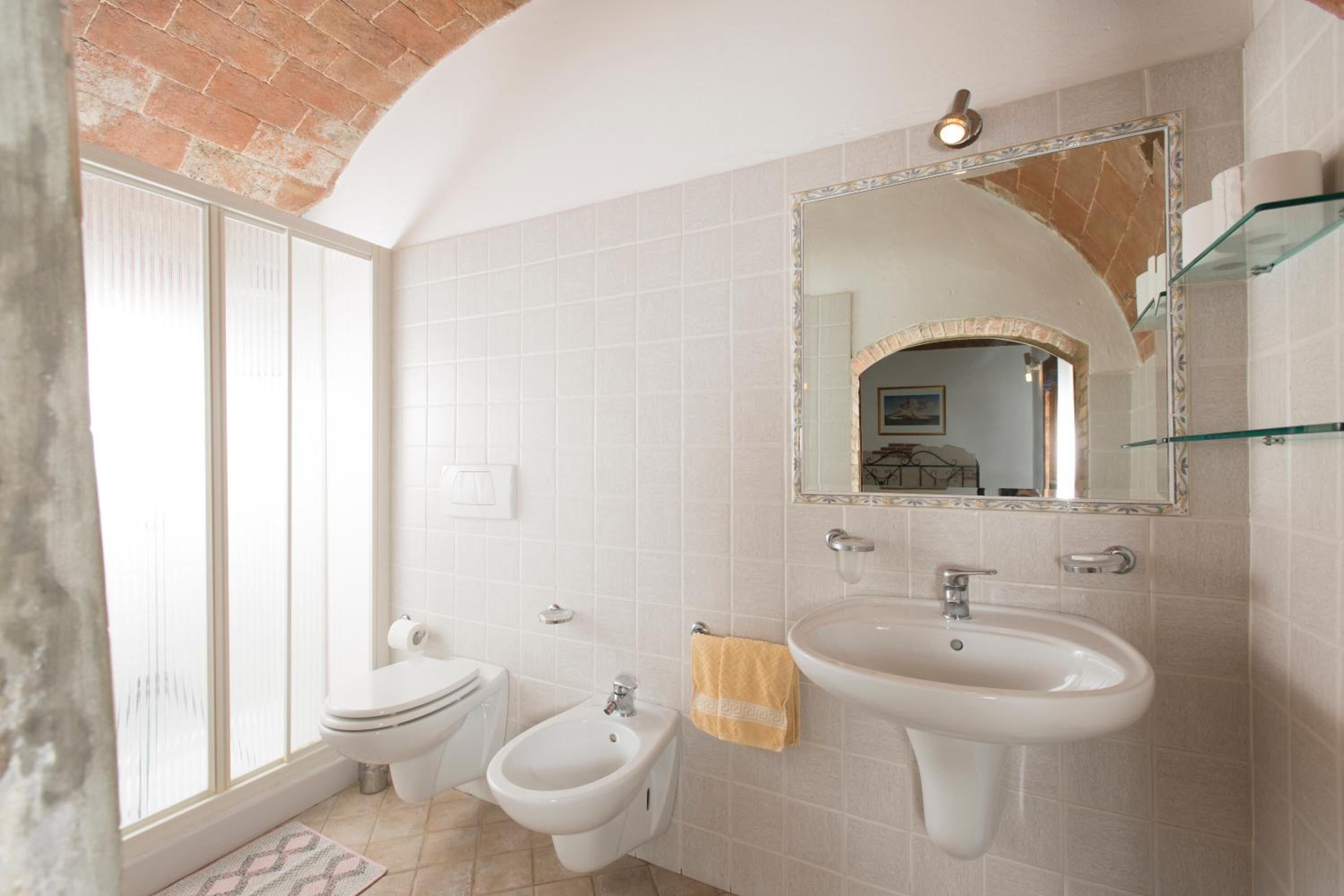 Un baño con inodoro, bidé y lavamanos blancos, un espejo grande y una repisa de cristal.