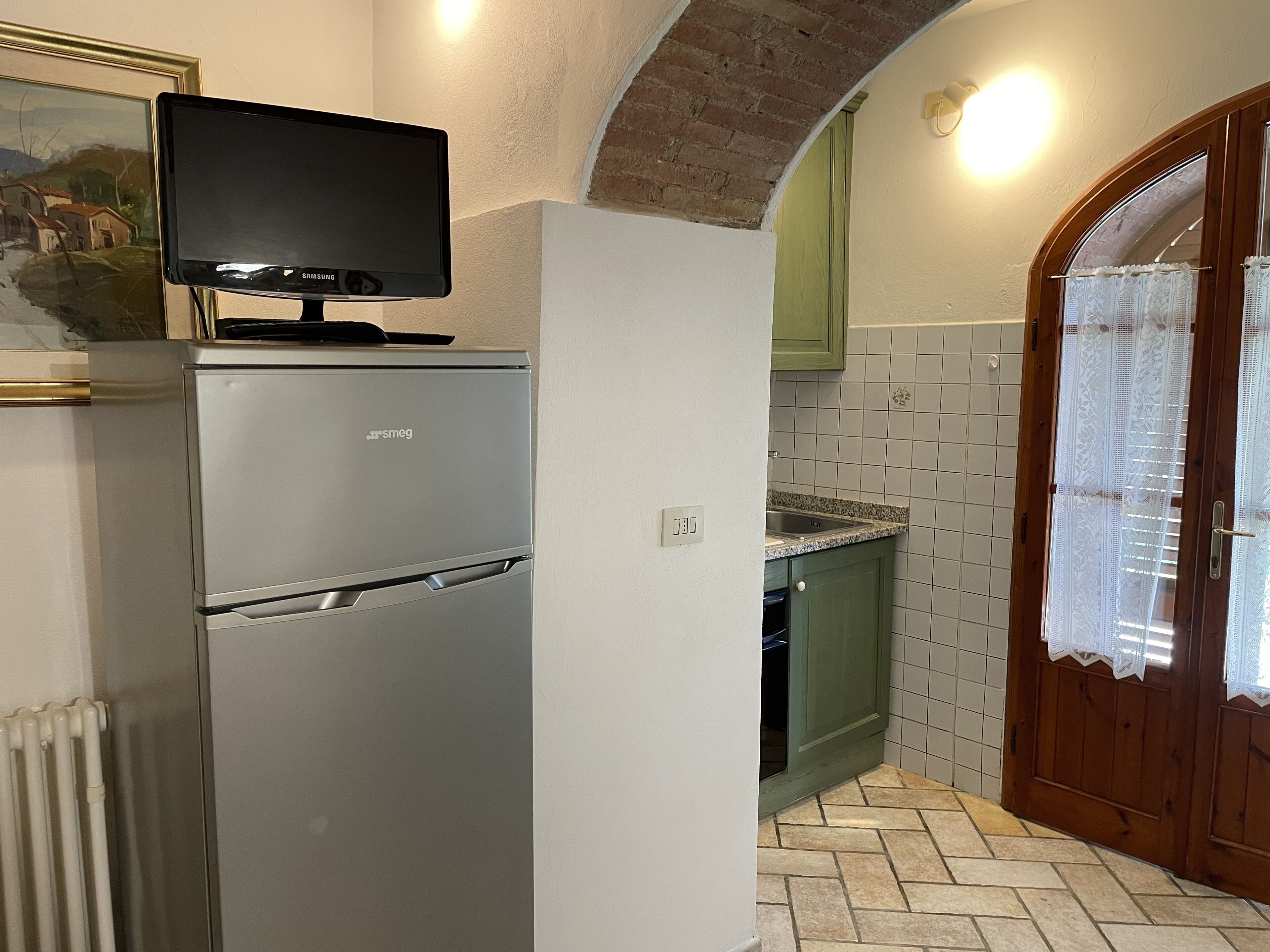 Een keuken met een koelkast, een televisie erboven, een betegelde spatwand en een houten deur met kantgordijnen.