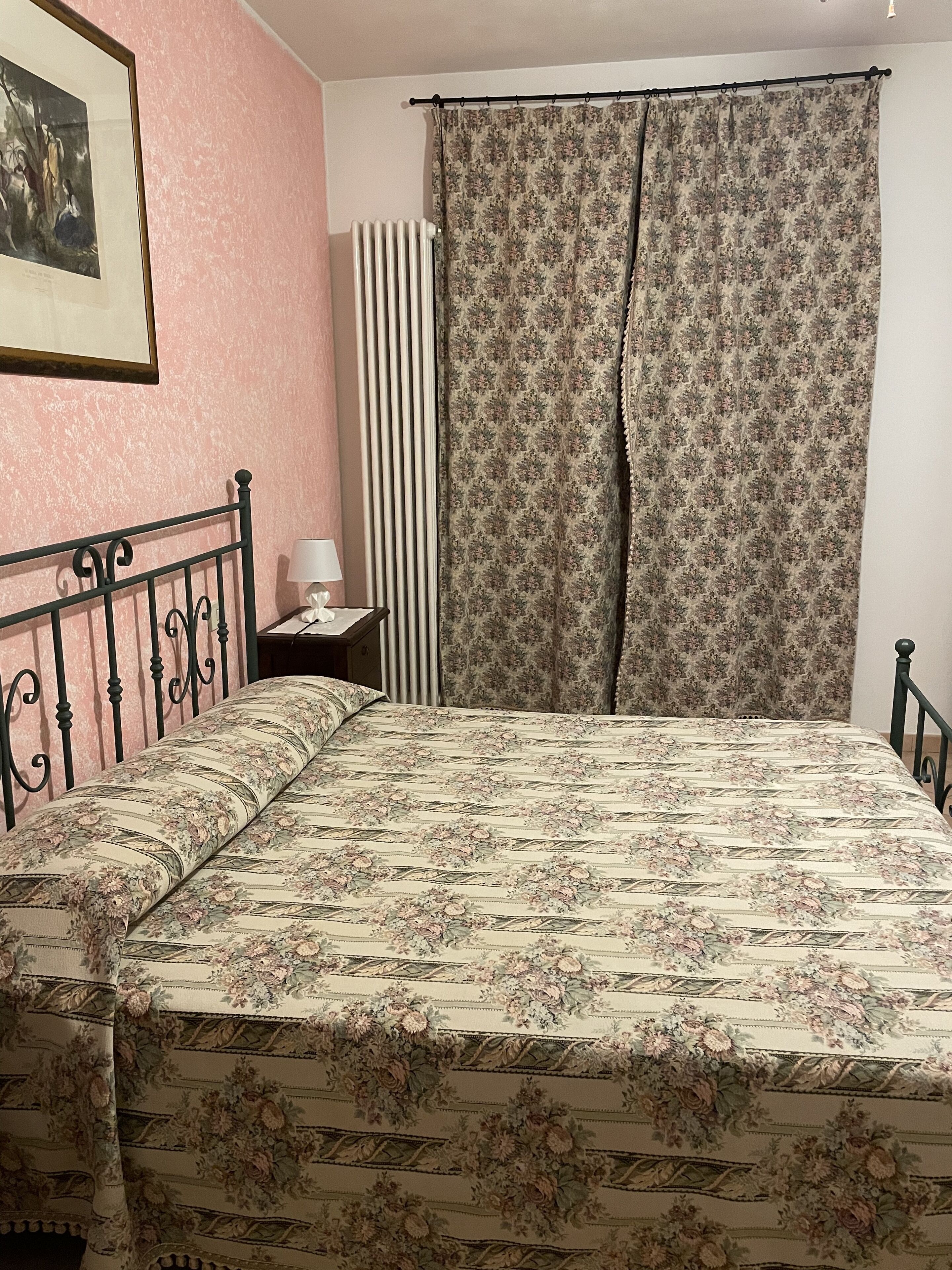 A bedroom with a bed, a nightstand, a lamp, a radiator, and a curtain.