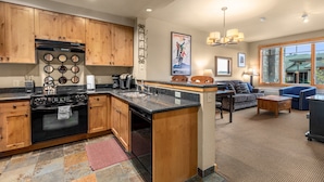 A modern kitchen with wooden cabinets, a black stove, and a large island. There is a living area with a sofa and a coffee table. - Incredible ski and summer condo!! Direct ski in and out, hot tub , gym, covered parking!! (Whitefish)