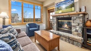 A cozy living room with a stone fireplace, a large window, and a television mounted above it. - Incredible ski and summer condo!! Direct ski in and out, hot tub , gym, covered parking!! (Whitefish)