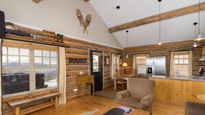 A log cabin interior with a wooden bench, a table with a skateboard, a sofa, a dining area with chairs, and a kitchen with a refrigerator. - Ski-in/ski-out "Alpine Escape" Cowboy Heaven Cabin-360 Degree Mtn View - Hot Tub (Big Sky)