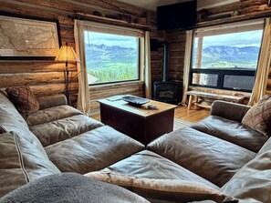 A log cabin interior with a leather sofa, a table, a lamp, a fireplace, and a large window with a mountain view. - Ski-in/Ski-out Cowboy Heaven Cabin - 360 Degree Mtn Views - Private Hot Tub (Big Sky)