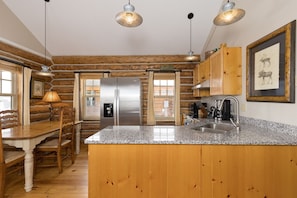 A log cabin interior with a kitchen area featuring a granite countertop, stainless steel appliances, wooden cabinets, and a dining table with chairs. - Ski-in/Ski-out Cowboy Heaven Cabin - 360 Degree Mtn Views - Private Hot Tub (Big Sky)