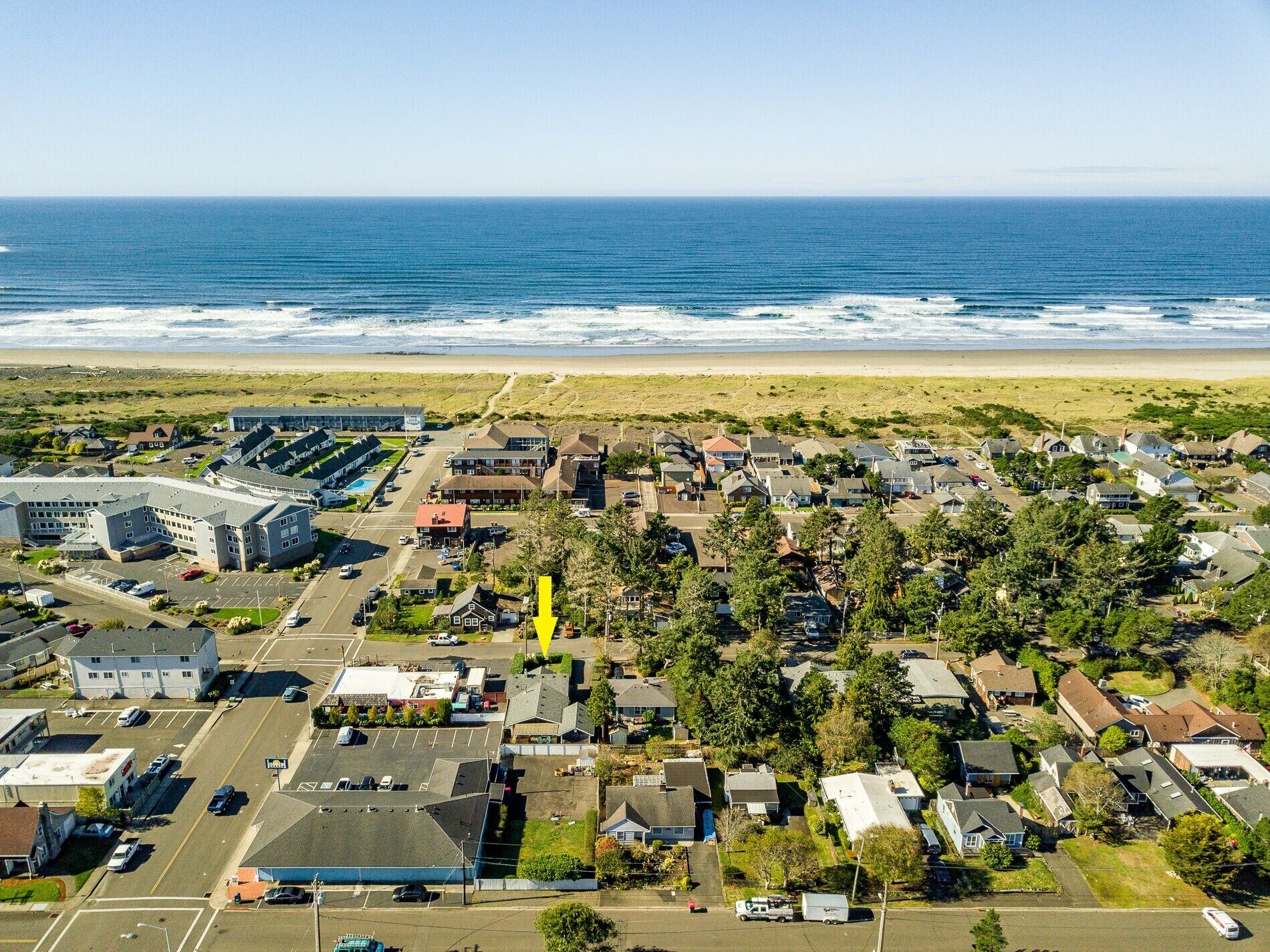 An aerial view of a coastal residential area with houses, a parking lot, and a beach in the background.