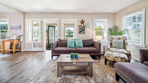 Un salon avec un canapé marron, une table basse en bois et un fauteuil à motifs floraux. Une porte donne sur un balcon, et une petite table est posée à côté, sur laquelle se trouvent un micro-ondes et une bouteille d’eau.
