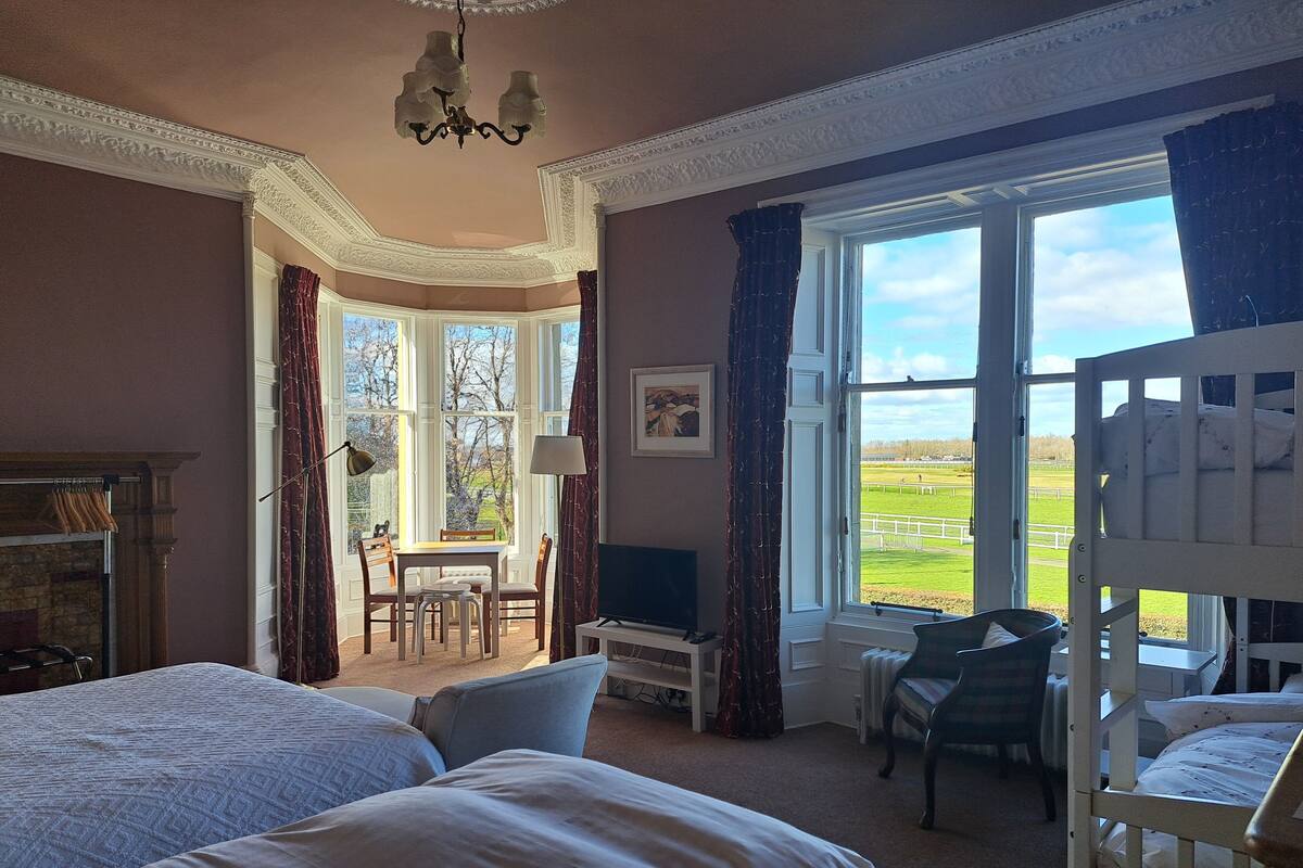 A bedroom with two beds, a desk, a chair, a television, and a view of a green landscape through large windows.