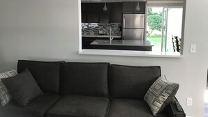 A modern living room with a dark grey sofa and patterned cushions, a built-in kitchen with stainless steel appliances, and a view of greenery outside.