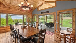 A wooden dining area with a large table and chairs, large windows with a view of the outdoors, and a chandelier.