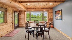 A wooden interior with a round table, chairs, and a large window offering a view of a green landscape.
