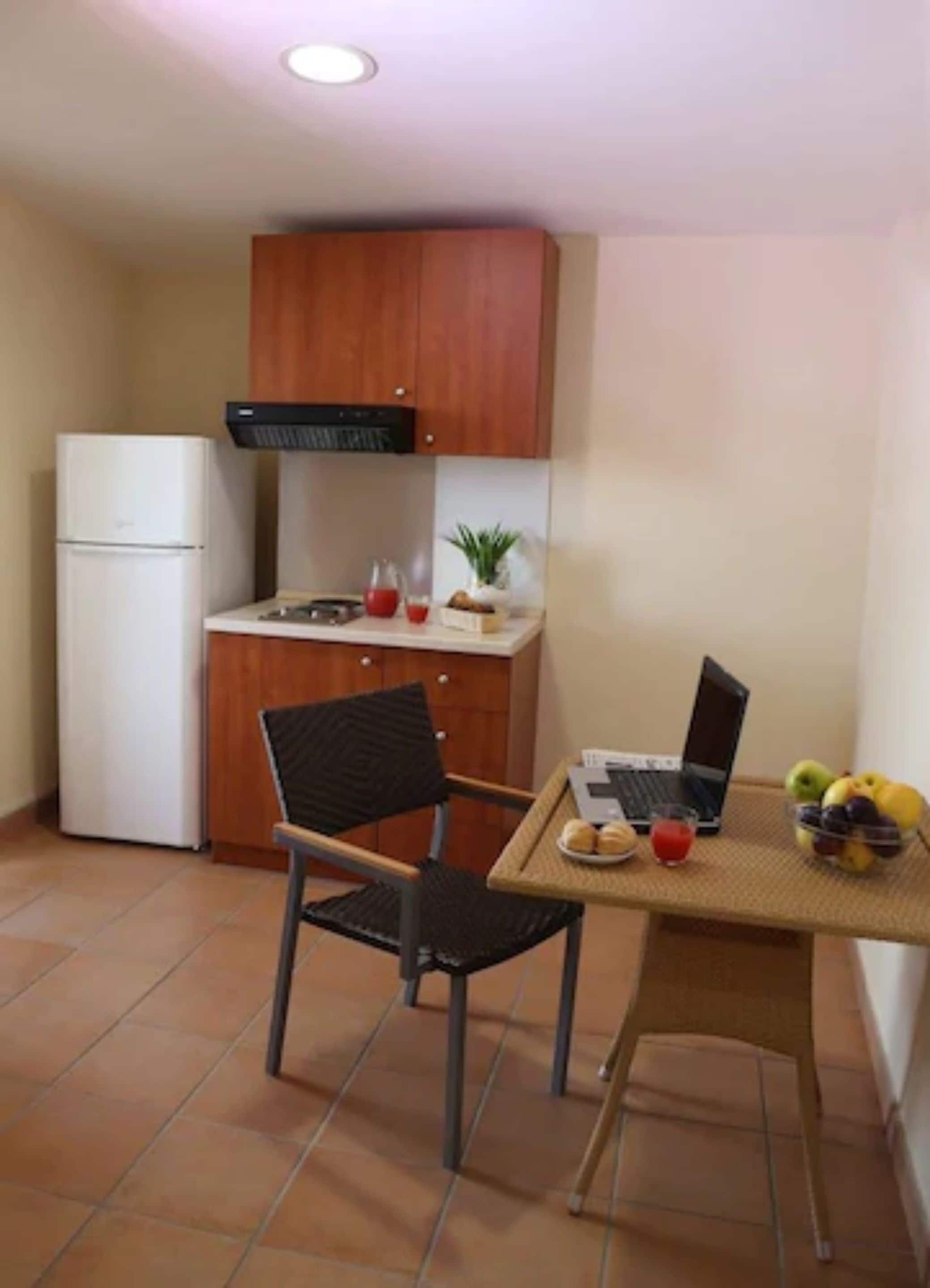 Een functionele keuken met een koelkast, een houten kast, een klein tafeltje met een laptop, een stoel en een schaal fruit.