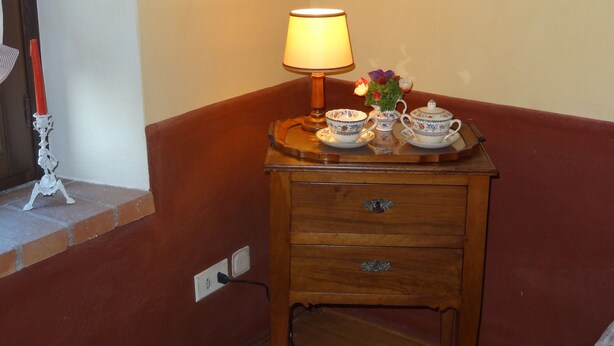 Una esquina de una habitación con una mesita de noche de madera que sostiene una lámpara, un florero con flores y un juego de tazas de té.