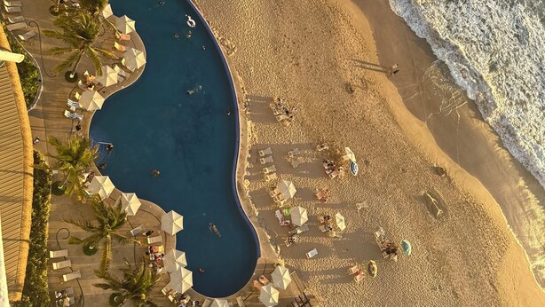 Vista aérea de una playa con piscina, sillas de descanso, sombrillas y palmeras.
