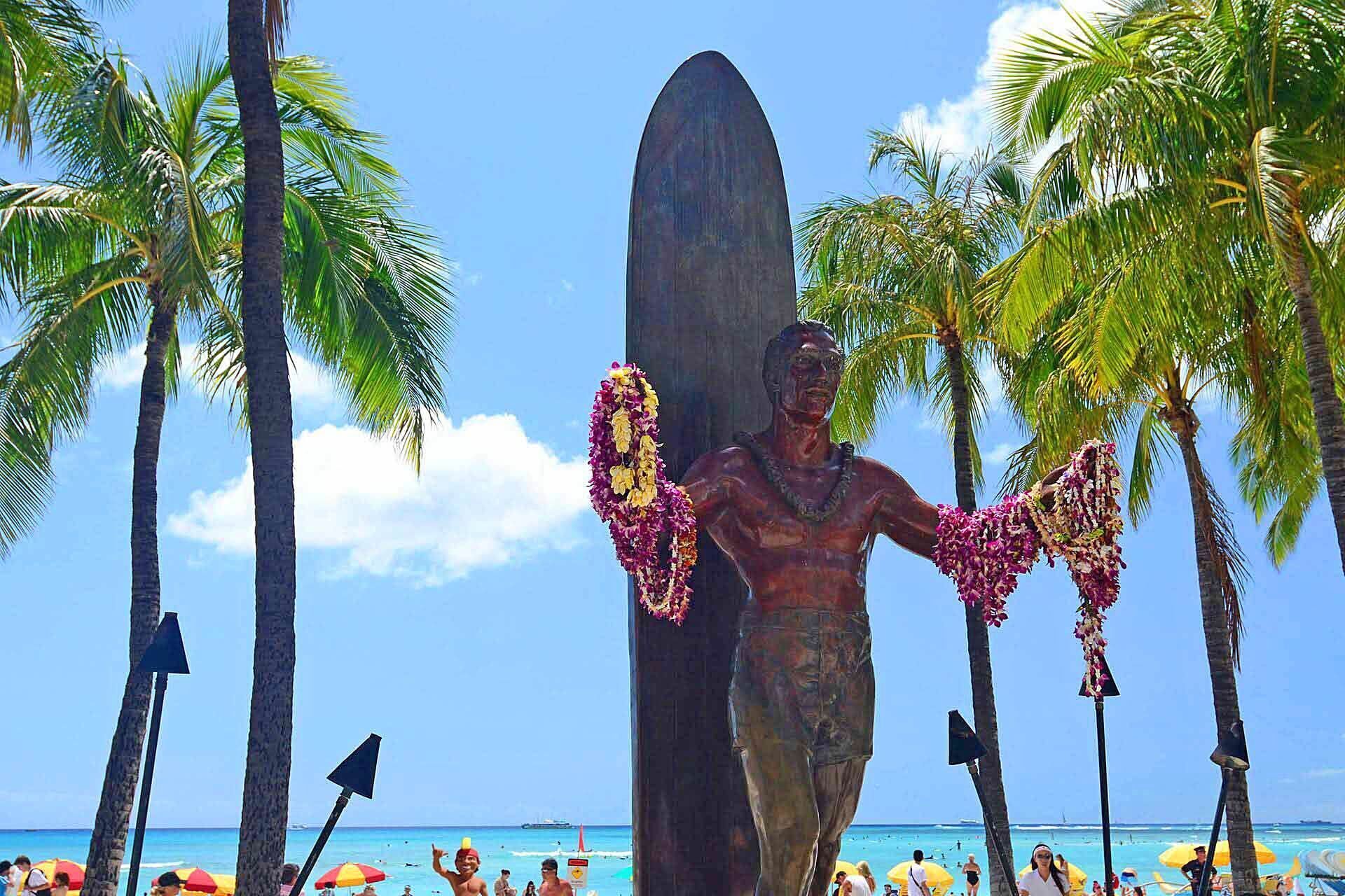 Eine Statue eines Mannes mit Surfbrett, umgeben von Palmen und Strandsonnenschirmen.