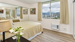 A hotel room with a bed, a nightstand, a mirror, a window with curtains, and a city view. - Great for Families! Partial Ocean/Canal/Diamond Head Views! Pool/WiFi/Parking (Honolulu)