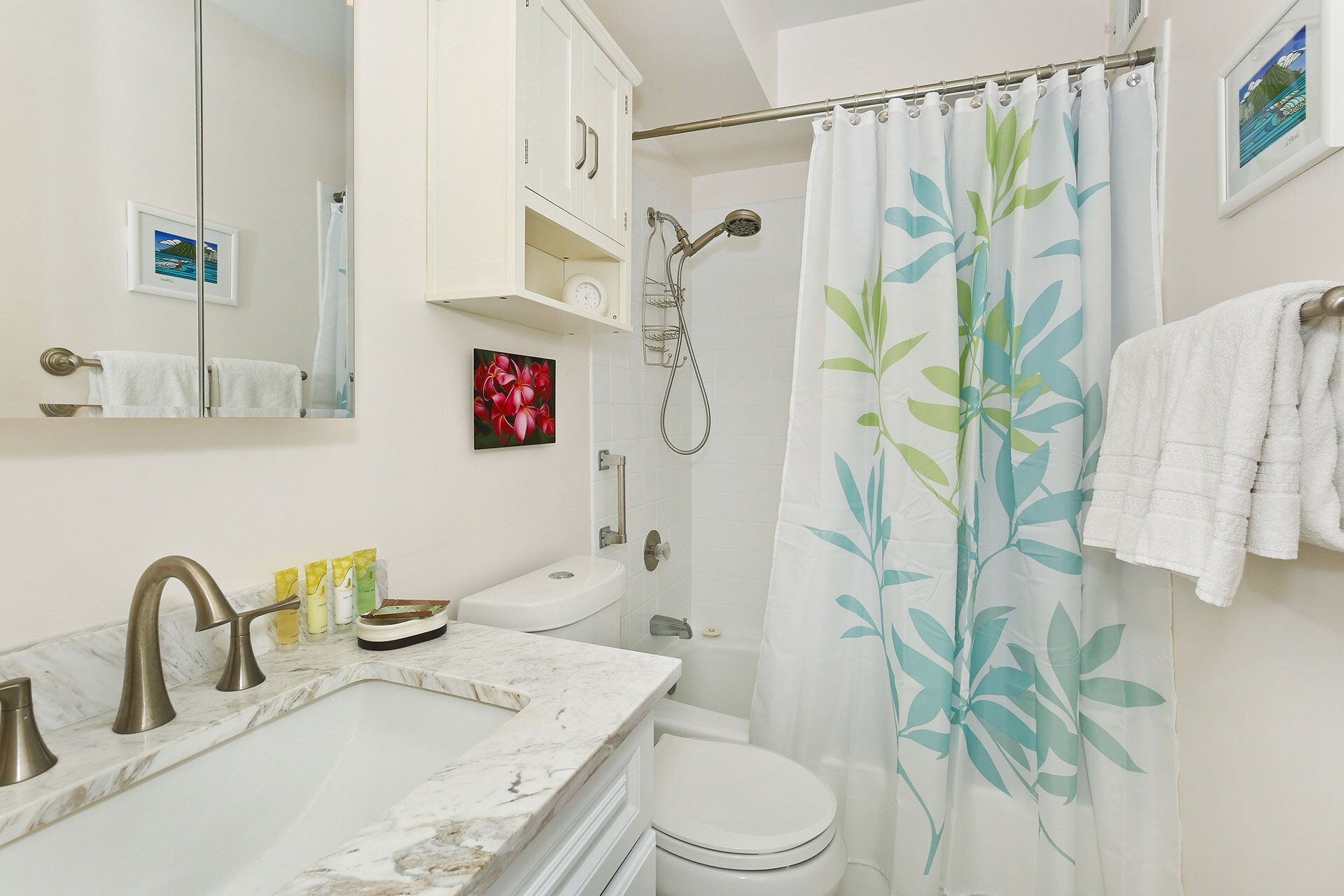 Une salle de bain avec un lavabo blanc, un comptoir en marbre, des toilettes, une douche avec un rideau à motifs de feuilles et un porte-serviettes.