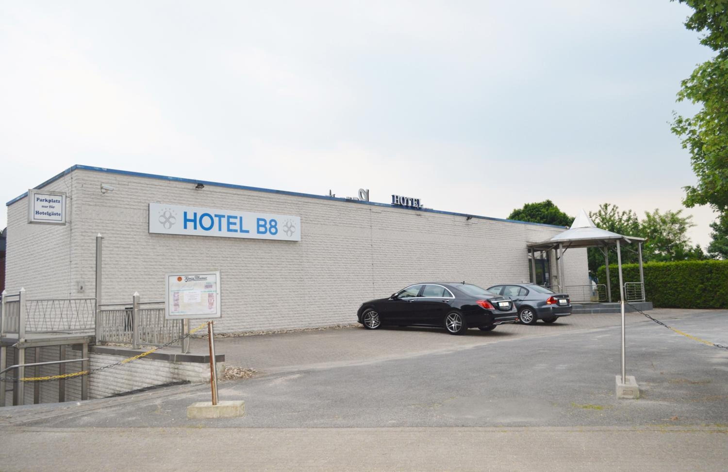 Un bâtiment d’hôtel avec une enseigne indiquant « HÔTEL B8 ». Deux voitures sont garées devant, et une zone abritée se trouve sur la droite.