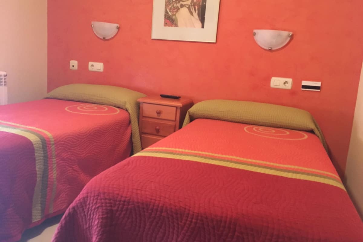 Une chambre avec deux lits, chacun recouvert d’une couverture rouge et agrémenté d’un oreiller vert. On y trouve une table de chevet avec une télécommande et un tiroir. Les murs sont peints en rouge, et deux appliques murales y sont fixées.