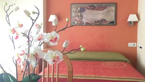 A room with a bed, a wall-mounted lamp, a painting, and a potted orchid.