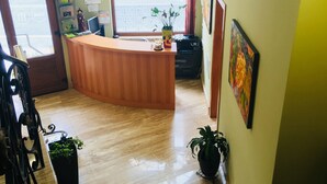 A reception desk with a wooden counter, a computer, and a potted plant. There are two potted plants on the floor and a painting on the wall.