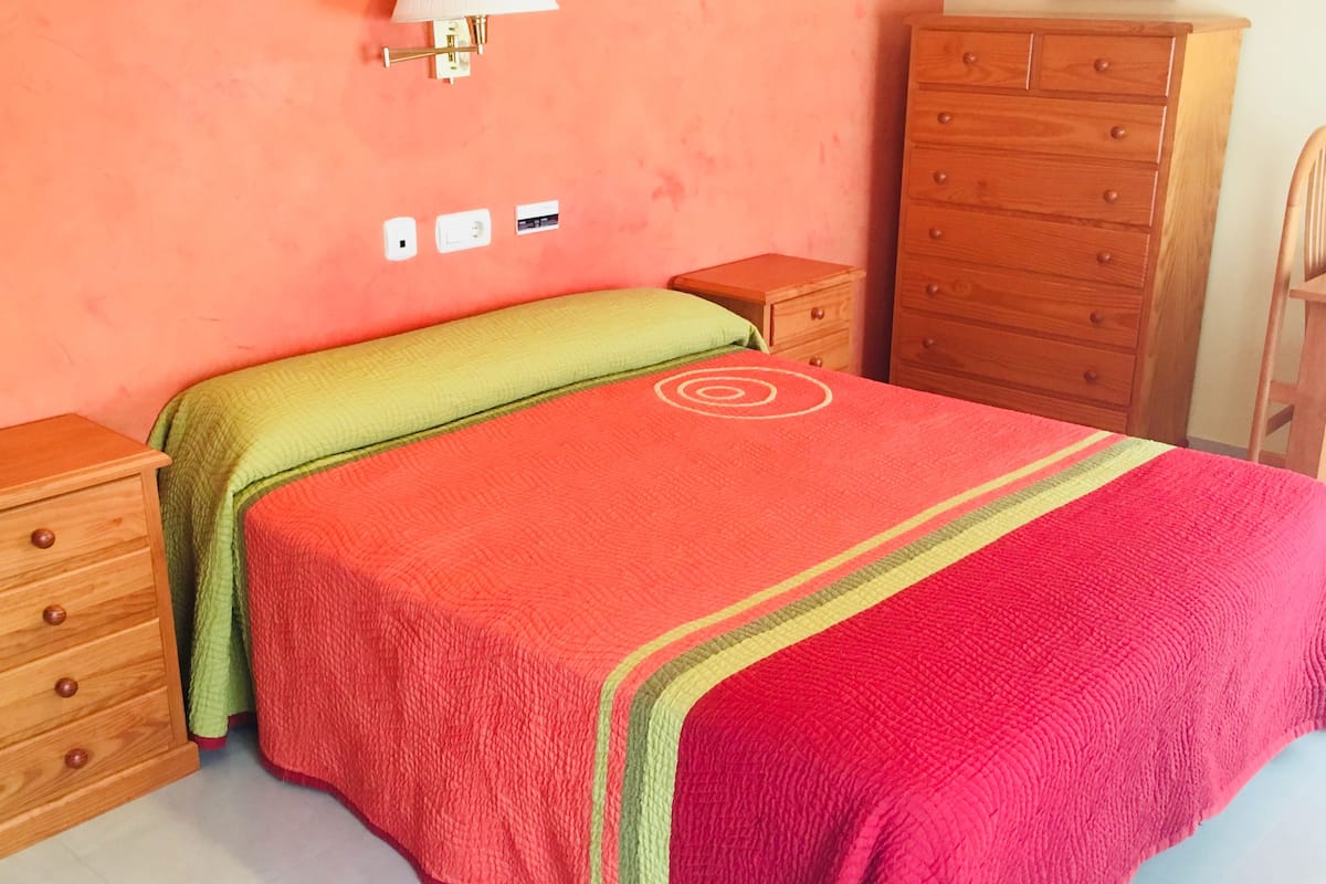 Une chambre à coucher comprenant un lit, une commode en bois, une table de chevet, une lampe et une prise électrique murale.