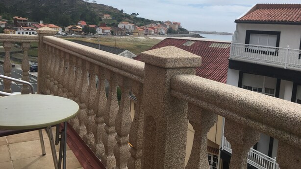 A balcony with a stone balustrade overlooking a coastal town with red-roofed buildings and a view of the sea.