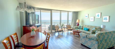 A spacious living area with a dining table, chairs, a sofa, and a balcony with a view of the sea.