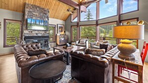 A spacious living room with a stone fireplace, leather furniture, large windows, and a ceiling fan.
