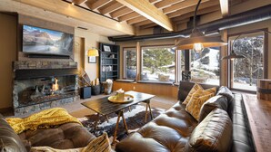 A cozy living room with a stone fireplace, leather sofa, and large windows offering a view of the outdoors.