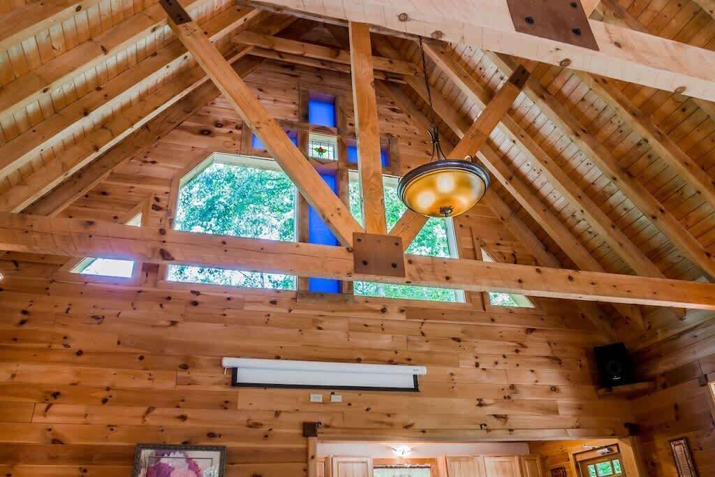 Interieur van een houten blokhut met een hoge nok, houten balken, een hanglamp en een raam met uitzicht op groen.