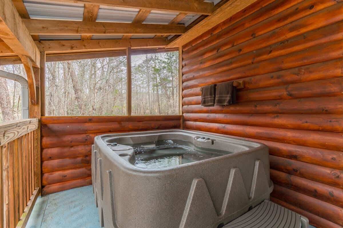 A log cabin interior with a hot tub, wooden walls, and a window overlooking trees.