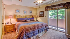A bedroom with a bed, two bedside lamps, a ceiling fan, and a window with a view of greenery.