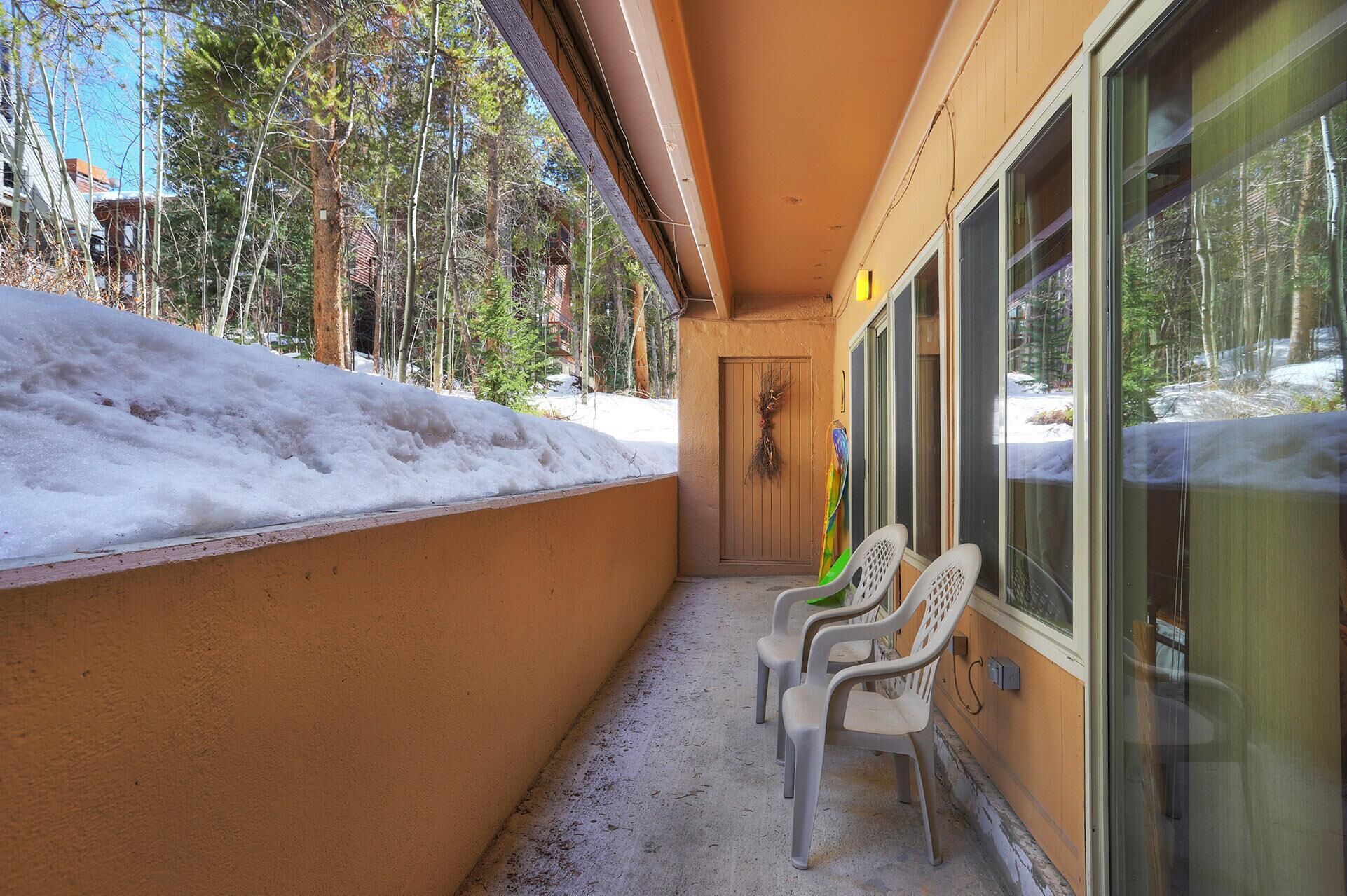 A covered outdoor area with a snow-covered wall, a door with a wreath, and three plastic chairs.