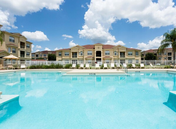 A swimming pool with lounge chairs and umbrellas, surrounded by a white fence and a row of buildings with a red roof. - Kinsale (Davenport)