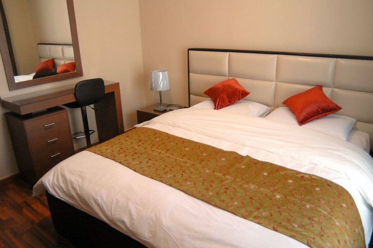 A hotel room with a bed, a desk with drawers, a chair, a nightstand, a lamp, and a mirror.