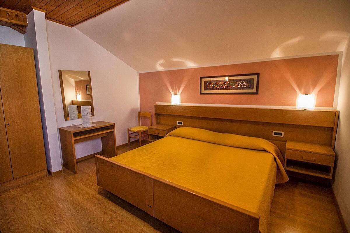 A bedroom with a wooden bed, a desk, a chair, a nightstand, and a wall-mounted lamp.