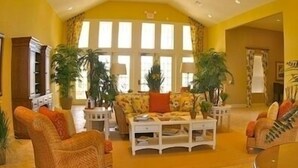 Amplia sala de estar con sofá, sillones y una mesa de centro. Se observan ventanas grandes con cortinas, plantas en macetas y un televisor.